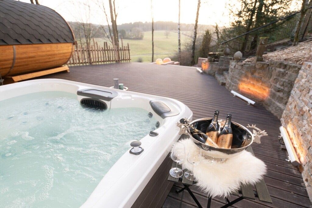 Ein Whirlpool mit Champagnerflaschen und Gläsern auf einer Holzterrasse, mit einer Steinmauer und Blick auf die Landschaft.