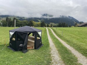 A tented outdoor area with a gravel path leading to it, surrounded by green fields and mountains in the background. - Chalet Legi Ferienhaus, Dusche oder Bad, WC, 4 oder mehr Schlafräume Chalet Legi by Interhome (Adelboden)