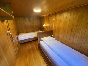A small, wooden room with two single beds, a nightstand with a lamp, and a ceiling light. - Chalet Legi Ferienhaus, Dusche oder Bad, WC, 4 oder mehr Schlafräume Chalet Legi by Interhome (Adelboden)