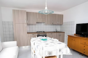Una cuina moderna amb armaris de fusta clara, una rebentada de rajoles blanques, una taula de dinar amb mantell blanc i un televisió de pantalla plana sobre un suport de fusta.