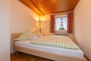 A bedroom with a wooden ceiling, a bed with a striped bedspread, a floor lamp, and a window with curtains. - Wank in the house of Georg (Garmisch-Partenkirchen)