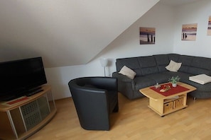 A living room with a television, a dark-colored sofa, a small black chair, a wooden coffee table with a red placemat, and a wall-mounted lamp. - Holiday apartment with 2 bedrooms (Wangerland)