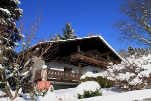 Een chaletachtige constructie met een houten balkon, omringd door met sneeuw bedekte bomen en een heldere blauwe lucht.