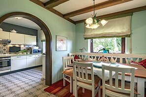 Ein Essbereich mit Holztisch und weißen Stühlen, eine Küche mit weißen Schränken und einer gefliesten Spritzschutzeinrichtung, und ein Fenster mit Blick auf Grünflächen.