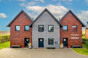 A modern brick building with a grey roof, two visible windows, and a door with a circular logo. - Meerhuus Carolinensiel House 1 (Carolinensiel)