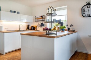 Eine moderne Küche mit weißen Schränken, einer Holz Arbeitsplatte, einem zweistöckigen Metallregal und einer Wanduhr.
