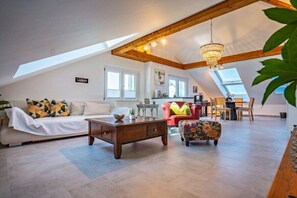 Un espaiós saló tipus loft amb un sostre alt, bigues de fusta, una pèndol, un sofà, una taula de centre i un pouf floral.