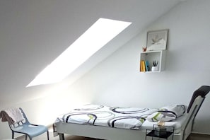 Ein kleines, modernes Schlafzimmer mit schrägen Wänden, einem Bett mit gemustertem Überwurf, einem Nachttisch mit Lampe, einem Sessel und einem Regal mit Büchern und Dekoration.