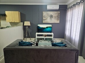 Ein Wohnzimmer mit grauem Sofa, einem Flachbildfernseher auf einem weißen Schrank, einer Lampe und einem Gemälde an der Wand.