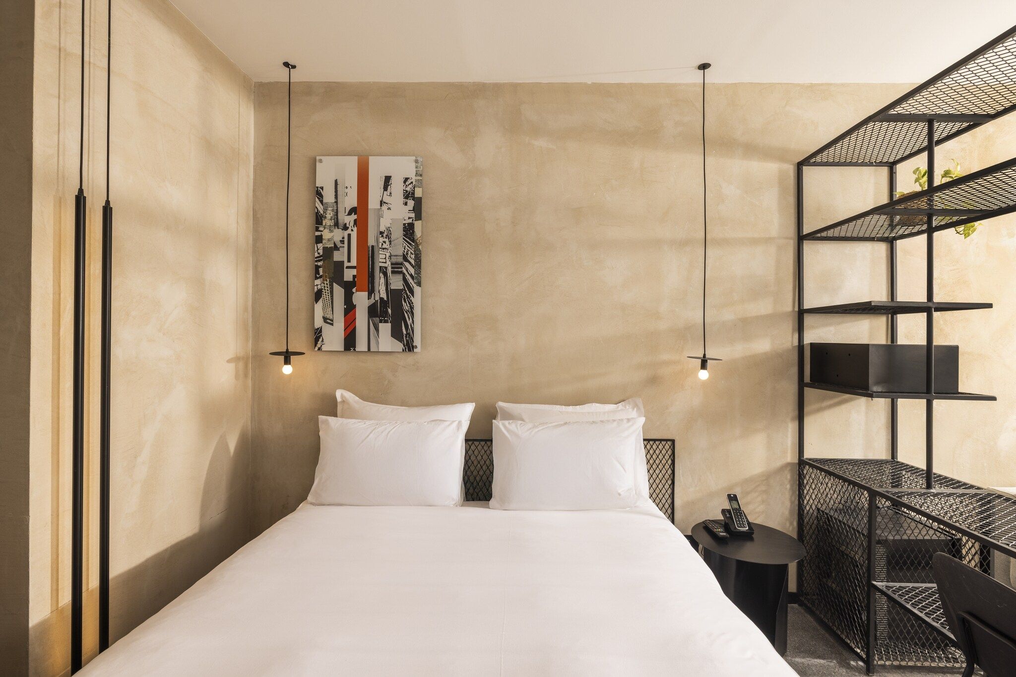 Quarto de hotel com uma cama, duas mesas de cabeceira, uma obra de arte na parede e uma estante de metal.