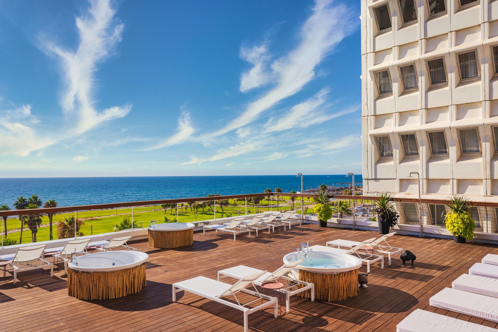 A rooftop terrace with lounge chairs, sun loungers, and hot tubs overlooking a beach and ocean.