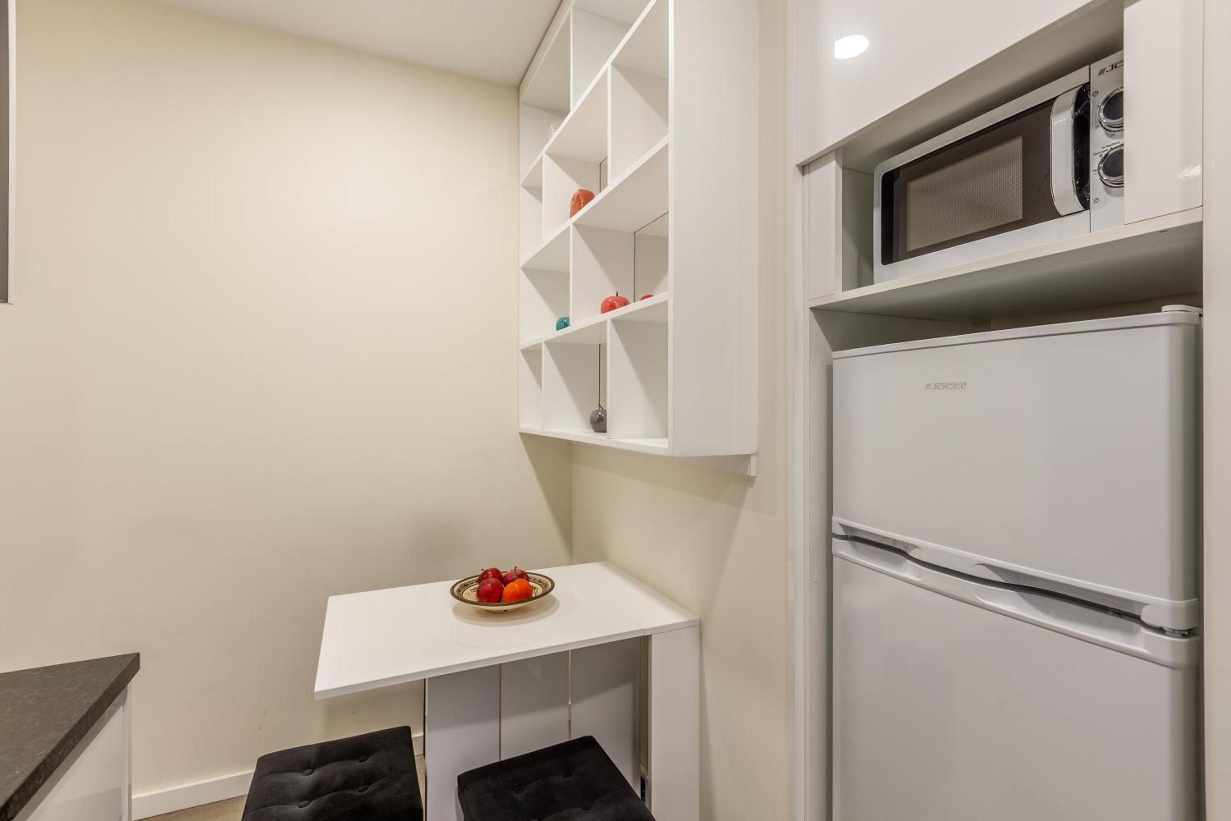 Een compacte kitchenette met een witte koelkast, een magnetron en een klein wit schapjesrek.