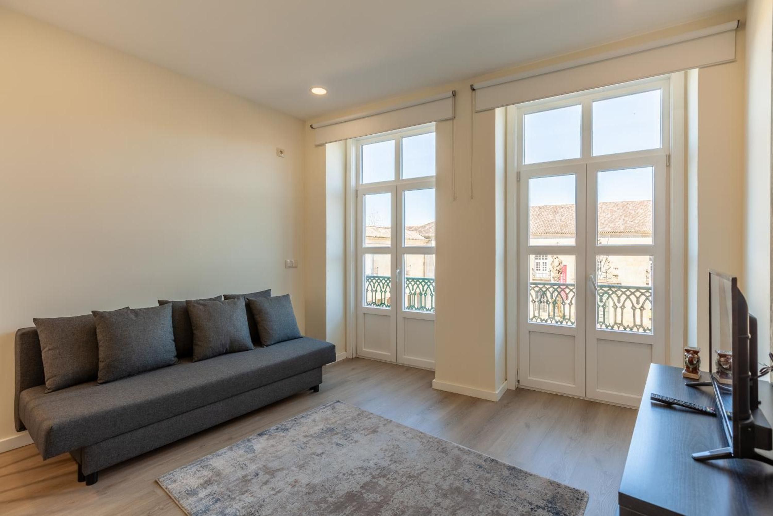 Una sala de estar con un sofá gris, un televisor de pantalla plana y ventanas amplias con vistas a un balcón.