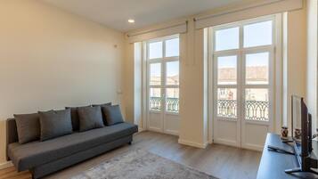 Una sala de estar con un sofá gris, un televisor de pantalla plana y ventanas amplias con vistas a un balcón.