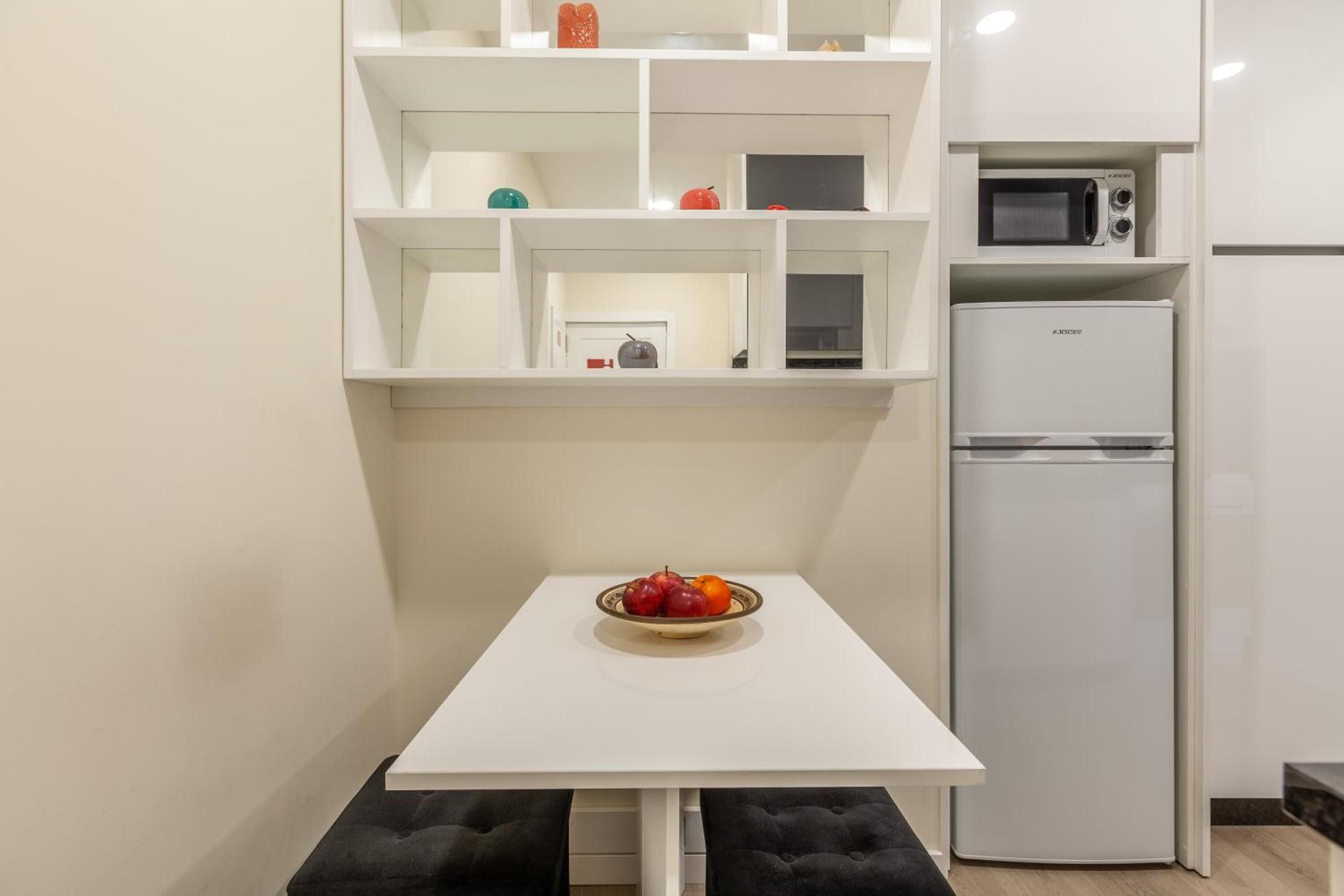 Una cocina pequeña con una mesa blanca, un tazón de fruta, un microondas y un refrigerador.