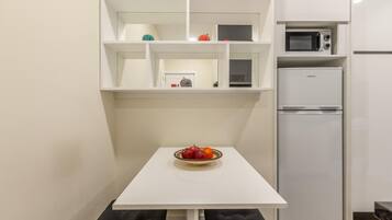 Una cocina pequeña con una mesa blanca, un tazón de fruta, un microondas y un refrigerador.