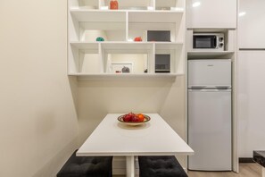 Una cocina pequeña con una mesa blanca, un tazón de fruta, un microondas y un refrigerador.