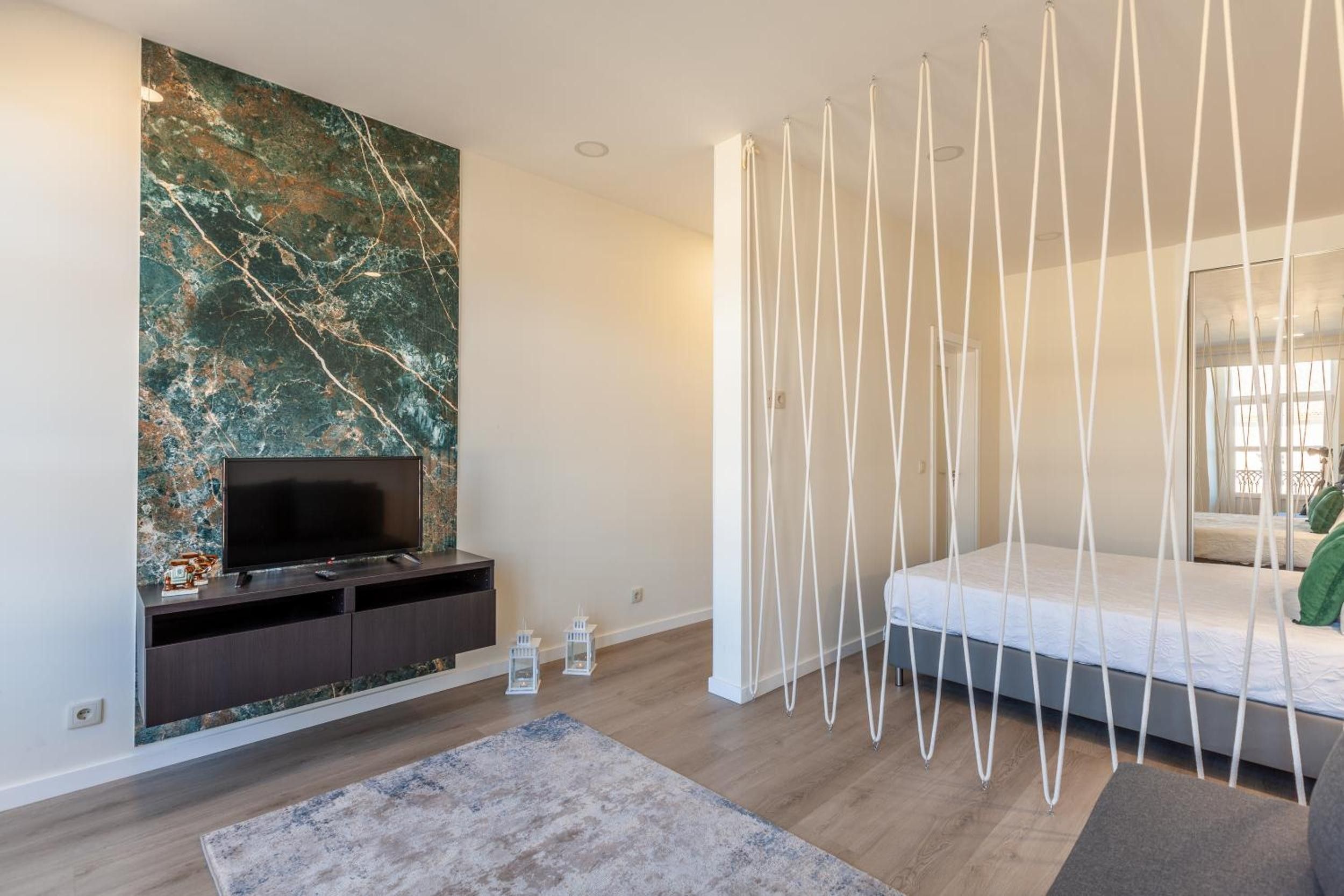 Un dormitorio moderno con una televisión sobre un mueble de madera oscura, una pared de mármol grande, una cama blanca con dosel y una alfombra gris.