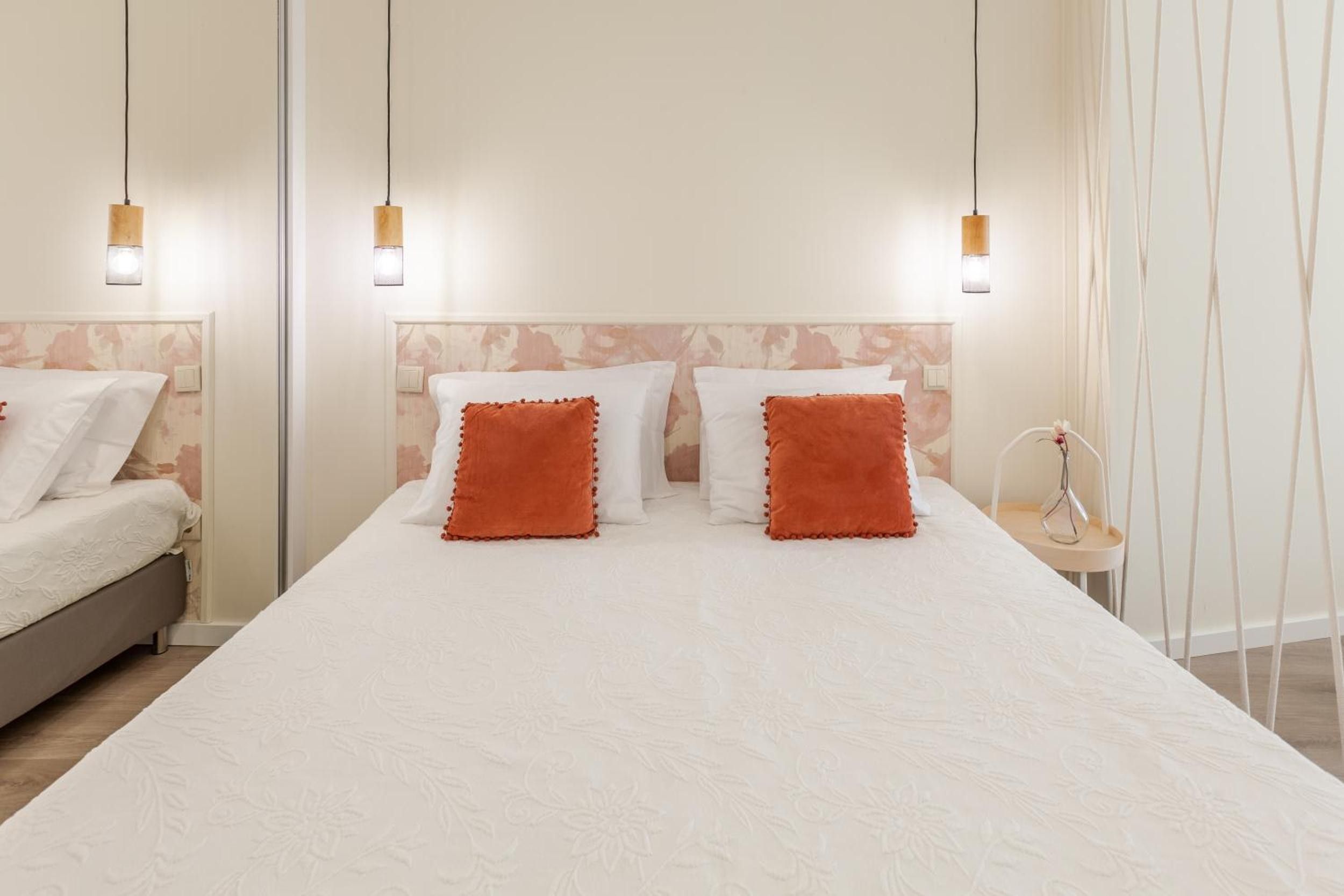 A double bed with white bedding and two orange pillows, a bedside table with a lamp, and a wall-mounted light fixture.
