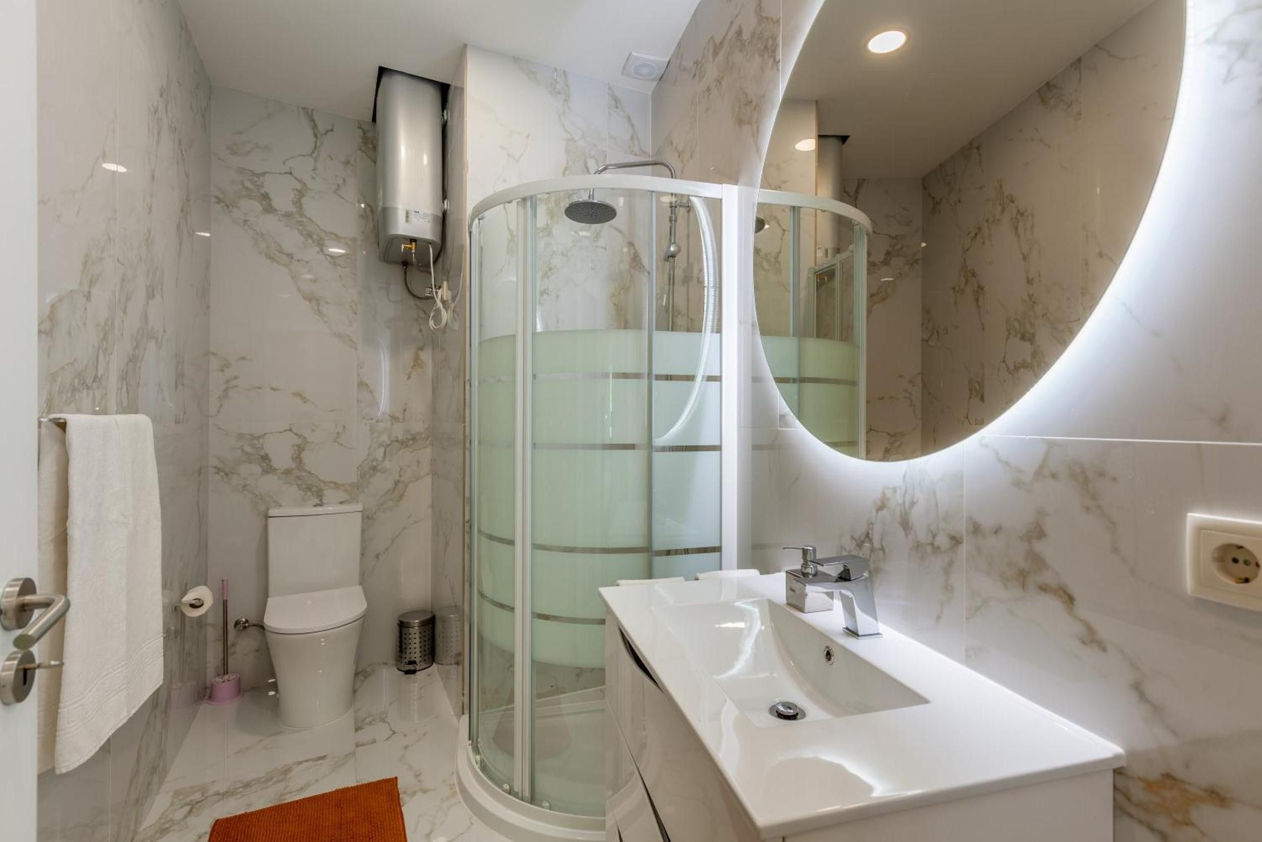 Un baño moderno con cabina de ducha de vidrio, un lavabo blanco y pared con azulejos de mármol.
