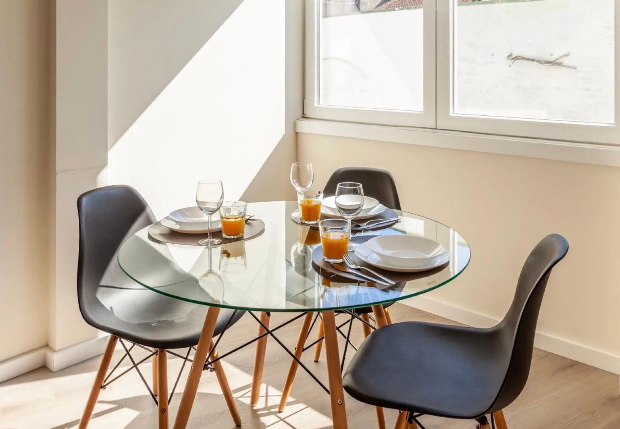 A modern dining area with a glass table and black chairs, featuring a breakfast setting with plates, glasses, and orange juice.