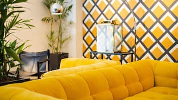 Um sofá amarelo em um quarto com parede estampada em padrão geométrico, uma planta em vaso e uma pequena mesa com abajur.