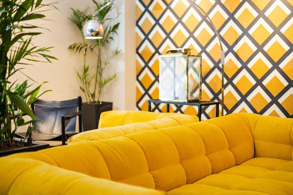 Un sofá amarillo en una habitación con un papel tapiz de estampado geométrico, una planta en maceta y una mesita con una lámpara.