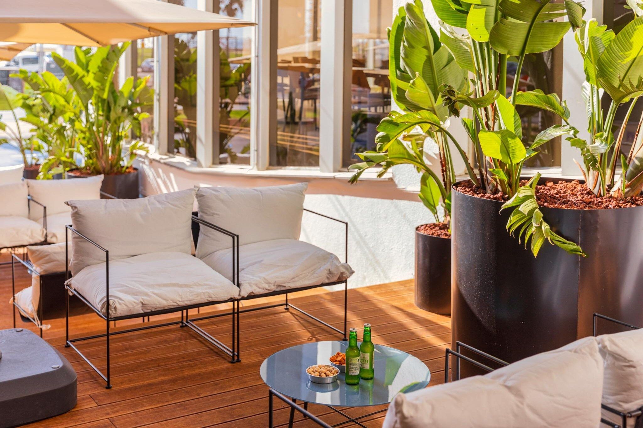 Un espace extérieur moderne, aménagé avec un canapé et des fauteuils, une table ronde ornée de bouteilles et de grands pots de plantes.