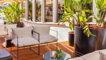 Un espace extérieur moderne, aménagé avec un canapé et des fauteuils, une table ronde ornée de bouteilles et de grands pots de plantes.