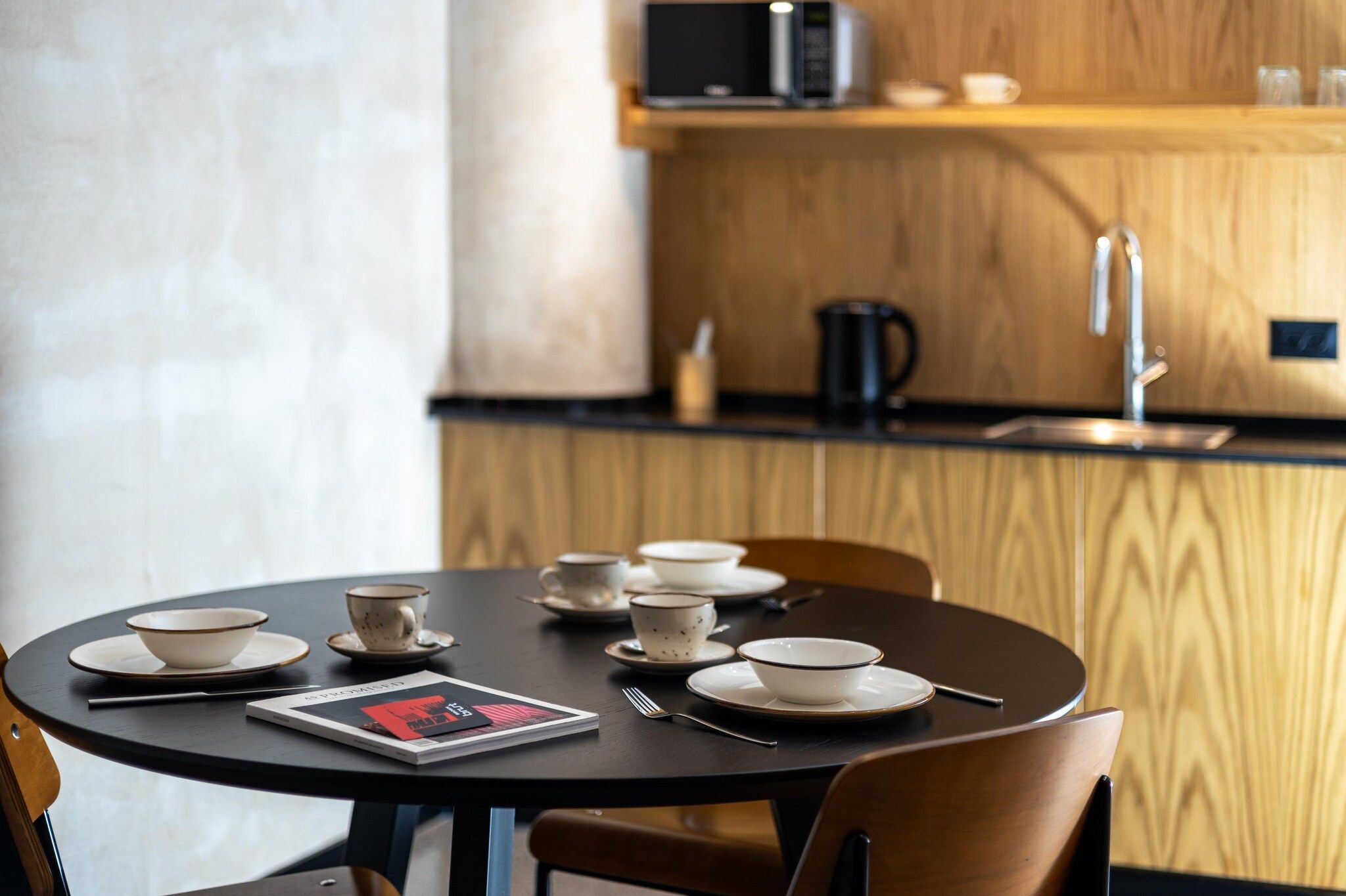 A round dining table with bowls, cups, and a magazine. A microwave and coffee maker are visible in the background.