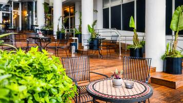 An indoor patio area with wooden flooring, metal chairs, and tables. There are potted plants and a large window in the background.