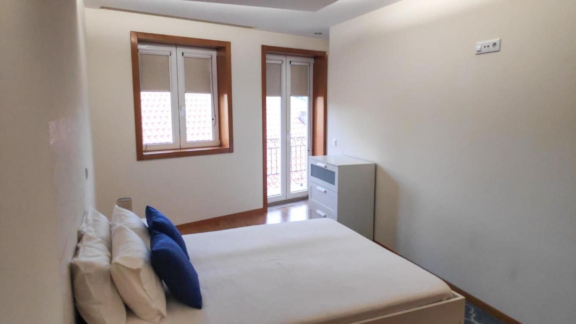 Un dormitorio con una cama, dos ventanas, un aparador y una puerta.