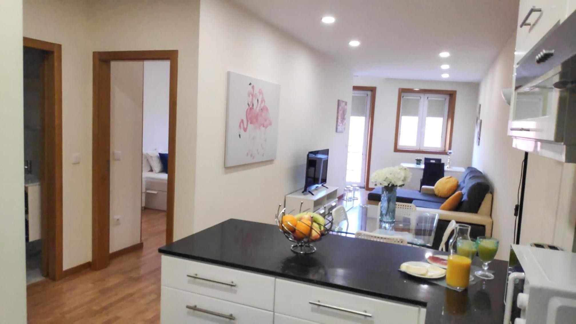 A modern kitchen with a black countertop, white cabinets, and a fruit bowl. There is a dining area with a sofa and a TV.