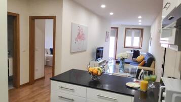 A modern kitchen with a black countertop, white cabinets, and a fruit bowl. There is a dining area with a sofa and a TV.