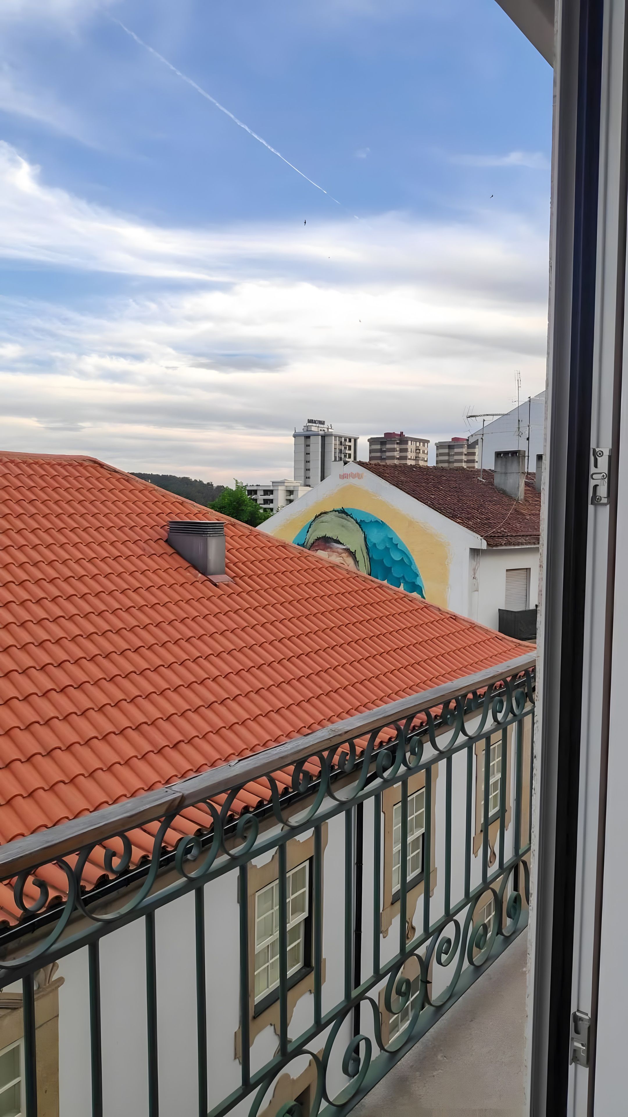 Un balcón con tejado de tejas rojas, barandilla de metal y vista a un edificio con un mural.