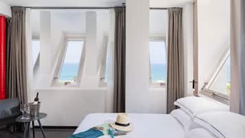 Habitación de hotel con una cama, una mesita con vasos y una botella, un sombrero y vista al mar a través de ventanas.