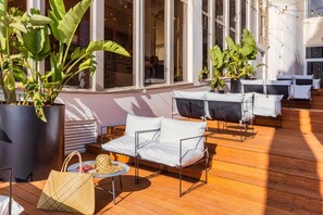 Een zonnig terras met een houten dek, witte gestoffeerde banken en plantenpotten.