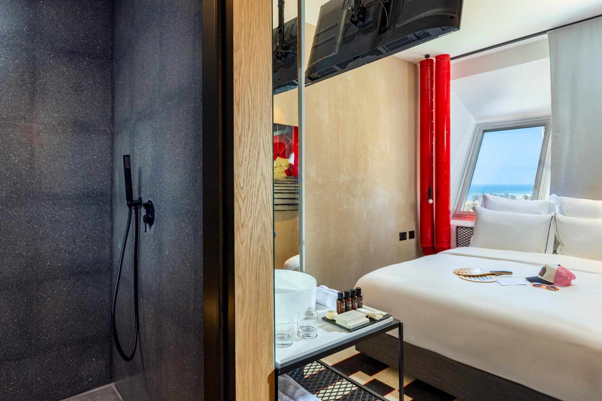 Quarto de hotel moderno com cama, mesinha, espelho e vista para o mar.