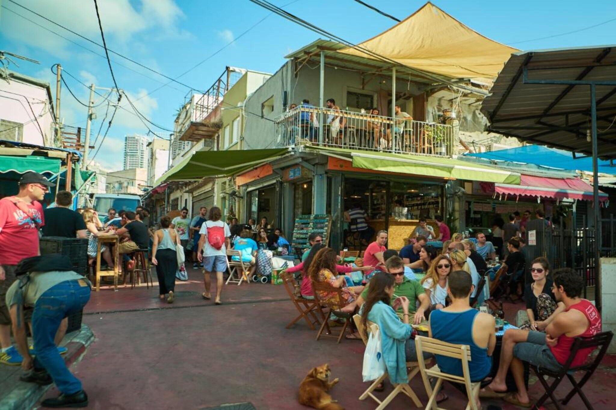 An outdoor dining area with tables and chairs, surrounded by buildings with balconies and awnings.