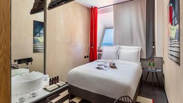 A hotel room with a bed, a bathroom with a sink and mirror, a small table with bottles, and a chair.