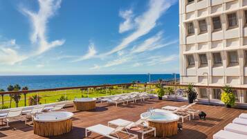 A rooftop terrace with lounge chairs, sun loungers, and hot tubs overlooking a beach and ocean.