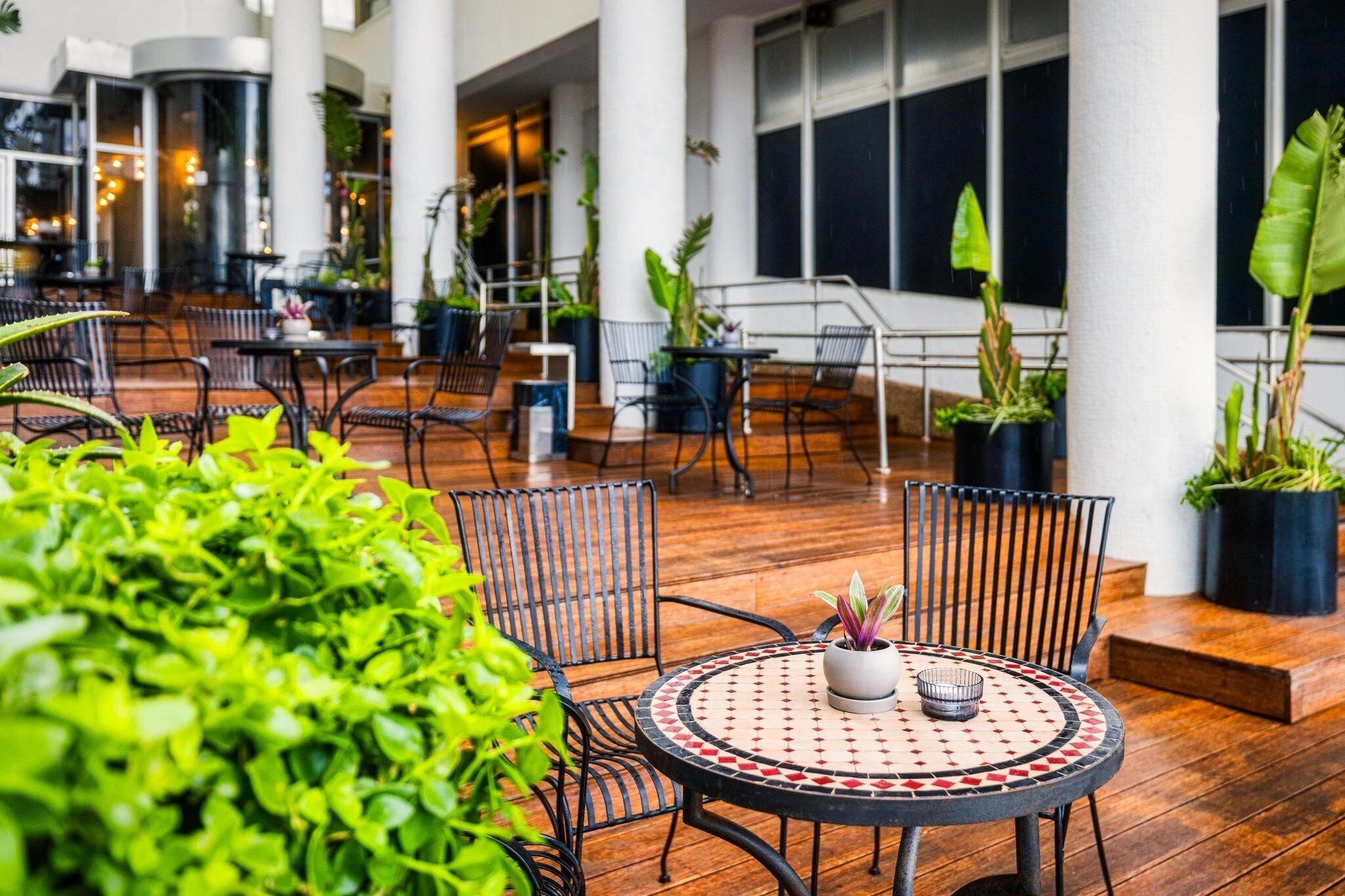 An indoor patio area with wooden flooring, metal chairs, and tables. There are potted plants and a large window in the background.