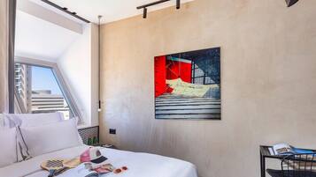 A hotel room with a bed, a window, a painting, and a small table with a magazine.