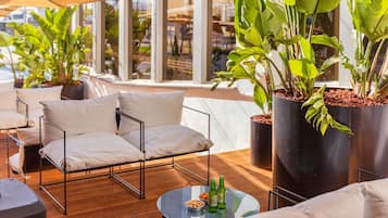 Uma área de estar moderna ao ar livre, com sofá e cadeiras, uma mesa redonda com garrafas e grandes vasos de plantas.