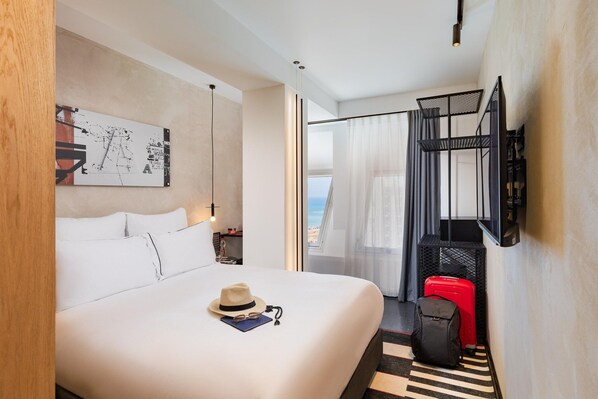 A hotel room with a bed, a desk, a chair, a suitcase, a hat, and a painting on the wall. - Dancenter Brut Seafront Hotel Tel Aviv- Standard Single Room (Tel Aviv)