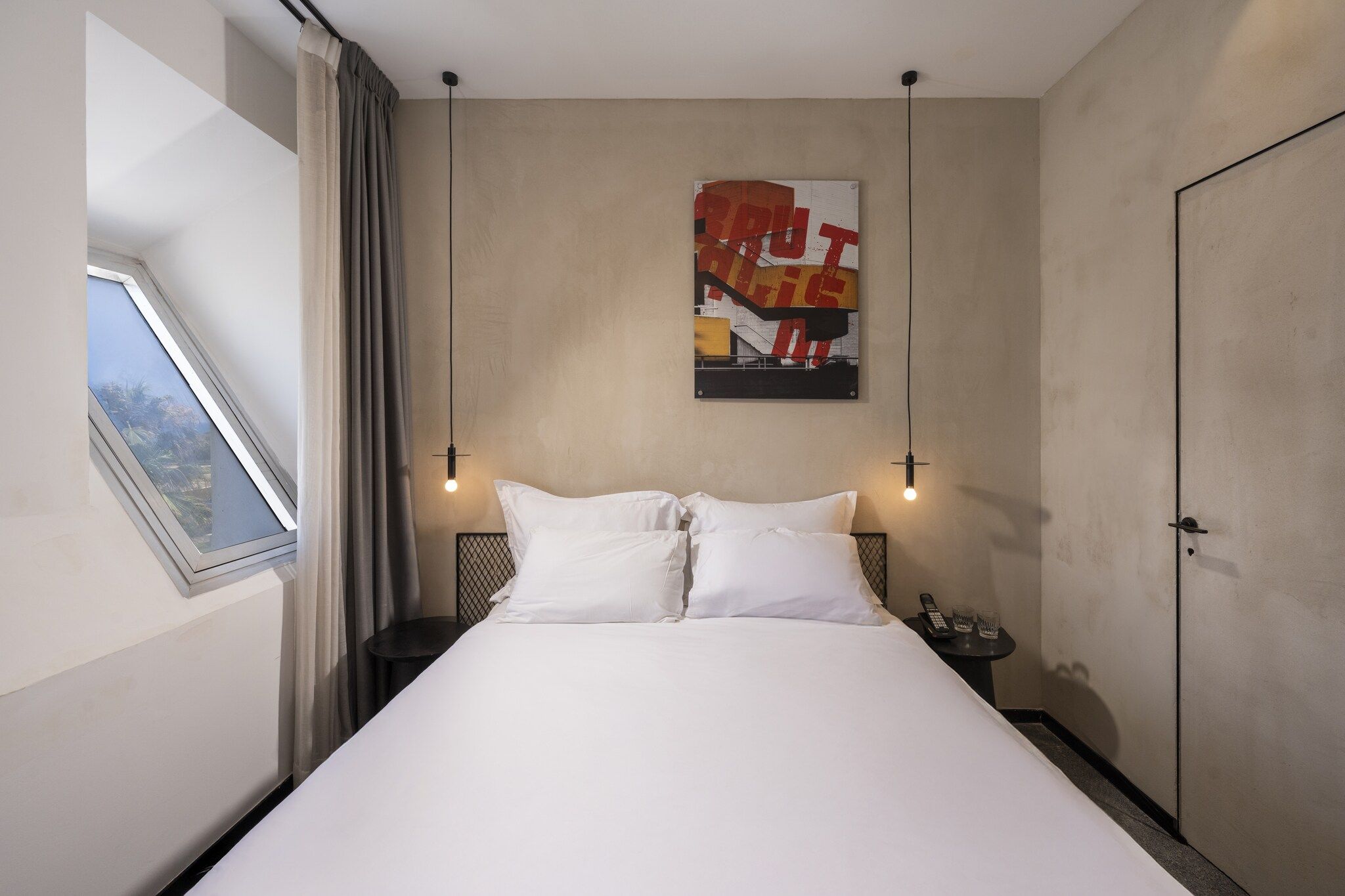 A hotel room with a bed, a window with curtains, a wall-mounted artwork, and a bedside table with a phone and glasses.