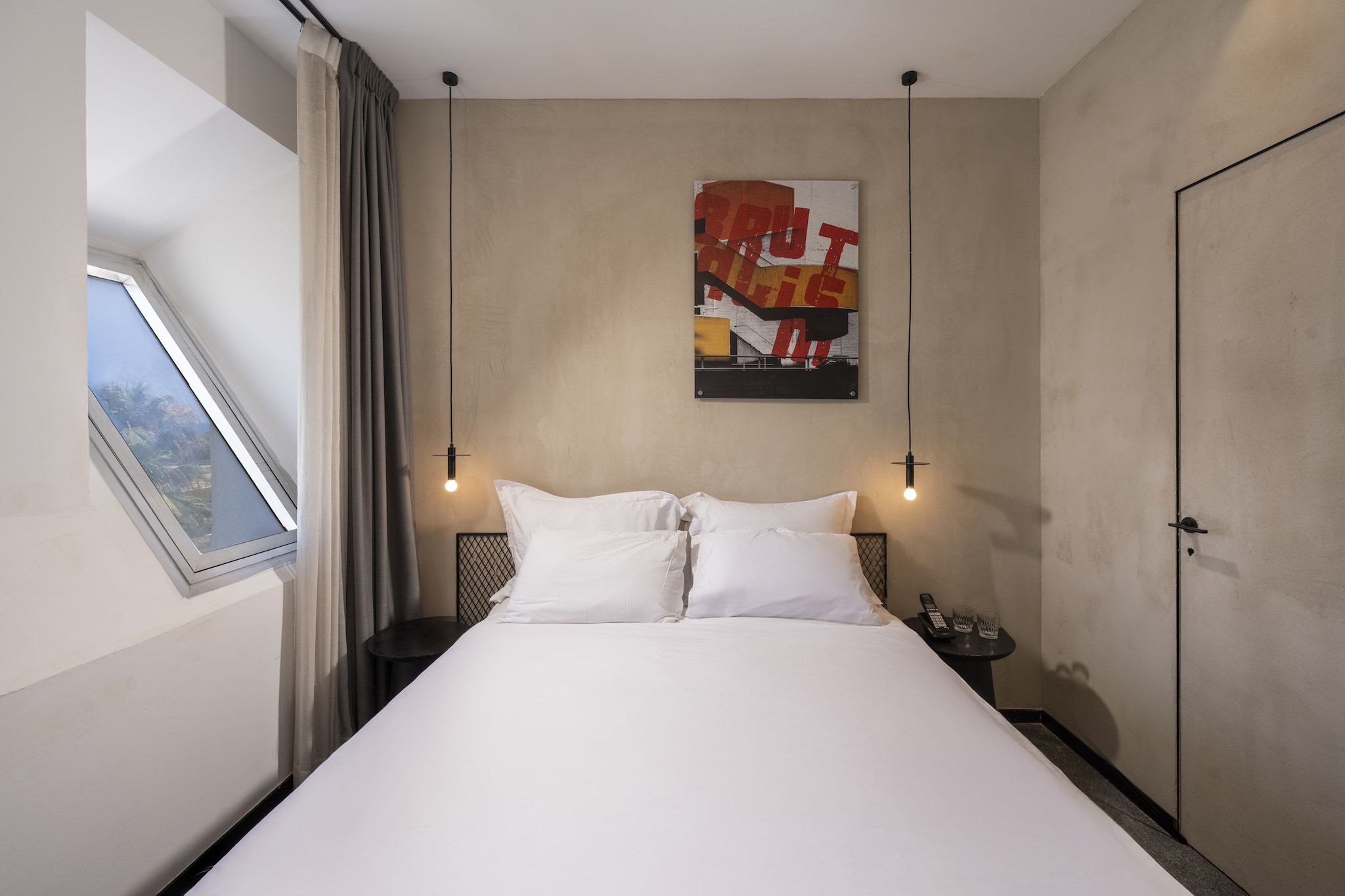 Habitación de hotel con cama, ventana con cortinas, una obra de arte colgada en la pared y una mesita de noche con teléfono y gafas.