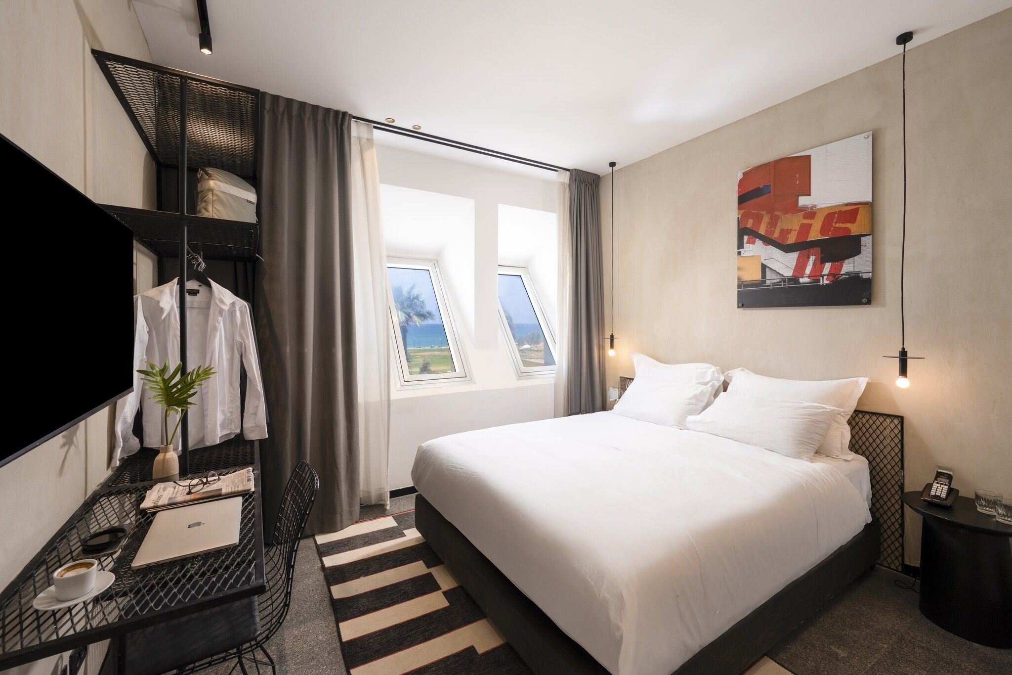 Une chambre d’hôtel avec un lit, un bureau avec un ordinateur portable, une chaise, une plante et une fenêtre donnant sur un paysage.