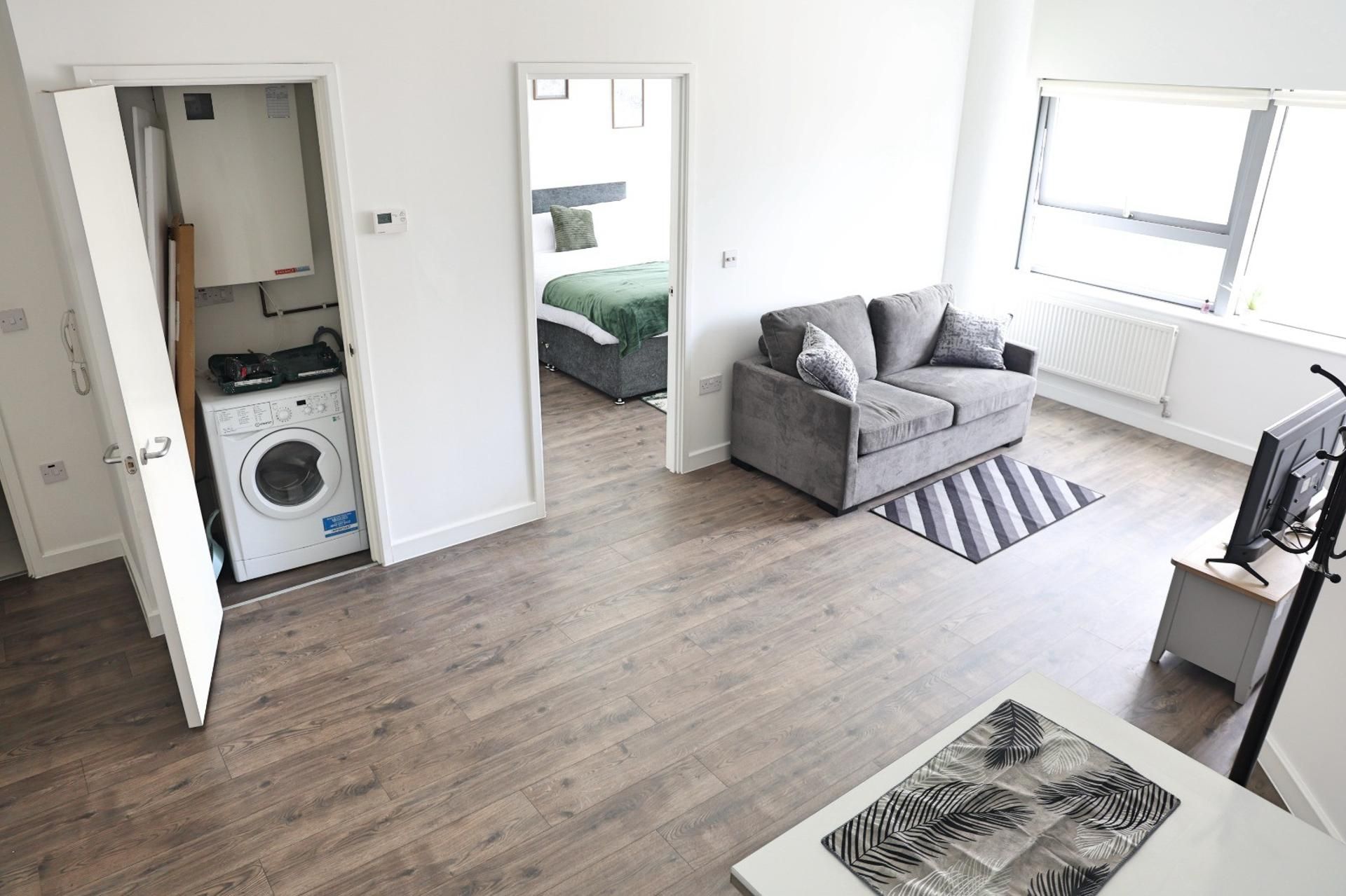 A modern apartment with a grey sofa, a flat-screen TV, and a washing machine in the corner.