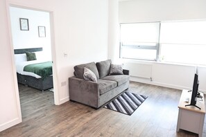 A modern living room with a grey sofa, a small rug, a wooden floor, a radiator, and a window. - Cozy Croydon Apartment Retreat (Croydon)